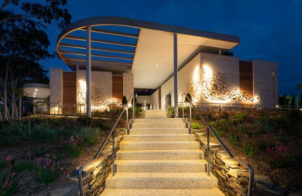 A dramatic dusk exterior view of a contemporary public or commercial building approached by a broad illuminated staircase. The building features a sweeping curved canopy with exposed horizontal slats, slim steel columns, pale masonry walls, timber-look cladding panels, and decorative organic illuminated wall graphics. Landscape planting and stacked-stone retaining walls frame the entry.