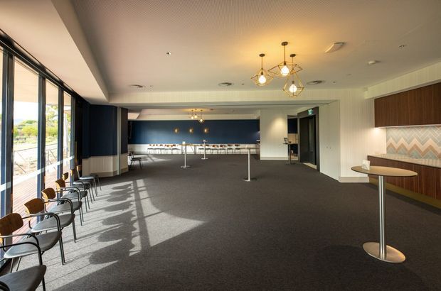 A spacious contemporary commercial lounge or hospitality area with floor-to-ceiling glazing along one side, dark carpet flooring, navy and white wall finishes, timber built-in banquette seating, small round standing tables, bar stools, and geometric pendant lighting. The room has a refined, warm-modern fit-out with strong natural light and a broad open circulation area.