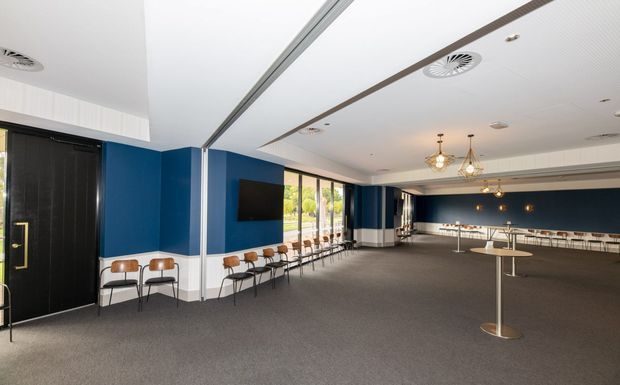 A large contemporary commercial interior with an open lounge or hospitality layout. Deep navy-blue wall sections are paired with crisp white ceiling and trim, dark doors, and a grey carpeted floor. Rows of small timber-and-black chairs line the walls, while slim pedestal tables occupy the open central area. A long counter or bar with stools is visible at the rear, beneath decorative pendant lights. Floor-to-ceiling glazing along one side brings in daylight and views of greenery outside.