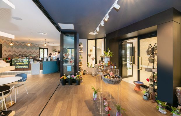 A contemporary café or retail hospitality interior viewed from the main customer area. The space combines a timber-look floor, dark charcoal walls and joinery, extensive glazing, track lighting, display shelving, indoor plants, and a colourful service counter and food display area. A patterned zigzag feature wall and small café tables and chairs are visible to the left, while retail-style displays and decorative objects occupy the foreground and right side.
