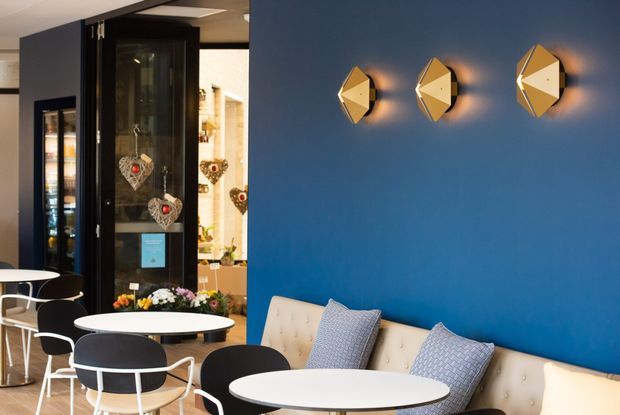 A contemporary commercial café or hospitality interior with a vivid blue feature wall, a long tufted cream banquette, small round white café tables, and black-and-white chairs. Geometric brass-toned wall lights create a decorative focal point, while a dark-framed glazed doorway opens toward a retail or display area. The space is bright, colourful, and polished.