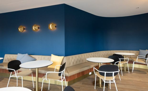 Contemporary lounge or café-style interior with deep navy-blue walls, a long built-in tufted beige banquette wrapping around the corner, small round white tables, and modern white-framed chairs with dark upholstered seats. Geometric brass wall sconces provide warm accent lighting, while timber-look flooring and a curved white ceiling create a polished, inviting setting.