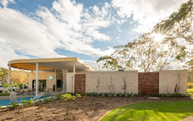Single-storey contemporary residential exterior with a broad flat-roofed covered outdoor area, pale rendered masonry walls, warm timber screening, and extensive glazing. A swimming pool and landscaped lawn are visible around the building, with mature trees and bright daylight creating an open suburban setting.
