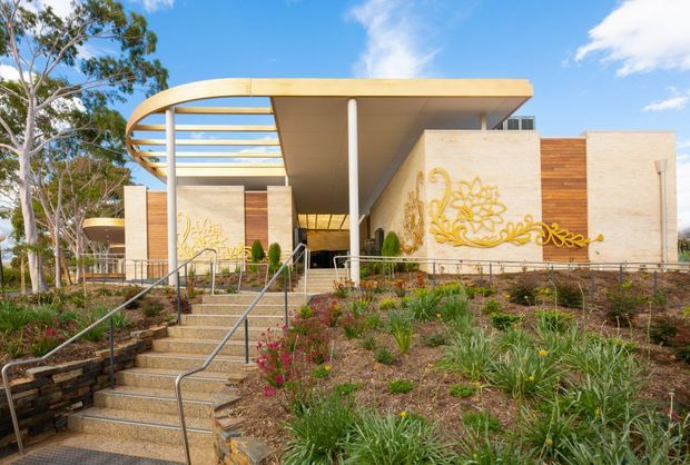 A contemporary low-rise building set within a landscaped sloping site, viewed from the front. The building features broad flat roof planes, a curved slatted roof canopy, exposed slender columns, pale rendered or masonry walls, warm timber-clad sections, and decorative gold-toned wall artwork. A central stair with metal handrails leads up through the planted garden to the entrance.
