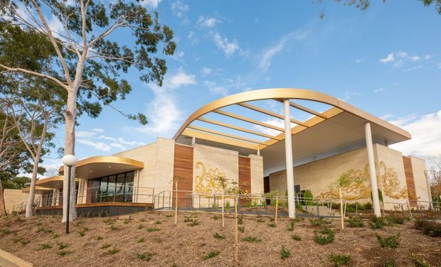 Exterior view of a contemporary low-rise building set among eucalyptus trees and newly landscaped grounds. The design features pale masonry walls, extensive glazing, warm timber-toned vertical cladding, curved roof canopies, slender white columns, and a large translucent-looking covered outdoor structure. Decorative mural-like artwork is visible on the facade.