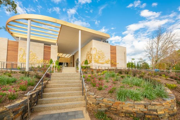 A contemporary public or institutional building viewed from landscaped grounds, featuring a broad entrance stairway, pale stone or concrete walls, warm timber-clad side walls, slender white columns, and a distinctive curved timber canopy with open slatted roof structure. Decorative gold-toned artwork is integrated into the entrance walls. Layered stone retaining walls and planted garden beds frame the approach under a bright blue sky.