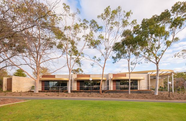 Single-storey contemporary institutional or commercial building viewed from landscaped grounds. The low horizontal facade uses warm tan masonry or rendered wall sections, dark glazed openings, timber-toned accent panels, and a prominent curved canopy at the right entrance. Mature eucalyptus trees, lawn, garden beds, and a paved approach frame the building.