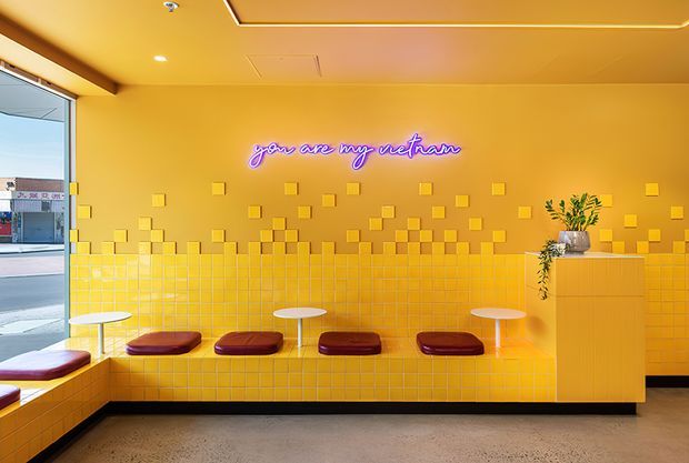 A vibrant yellow-orange commercial interior with a tiled built-in banquette, burgundy seat cushions, small round tables, and a yellow tiled wall featuring raised square tiles and a purple neon sign reading “you are my sunshine.” A large glazed opening reveals the street outside, while a plant sits on a tall yellow plinth. The space has integrated linear ceiling lighting and a polished concrete floor.