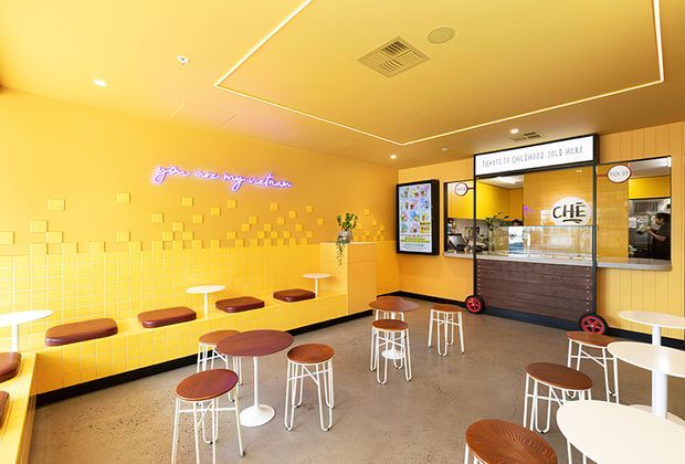 Bright, playful fast-food dessert or beverage shop interior with a yellow tiled feature wall, yellow ceiling and walls, neon wall lettering, scattered small square tile reliefs, compact café tables and stools, and a service counter with menu displays. The space has polished concrete flooring, white metal-framed stools, warm wood-toned tabletops, and a bold high-contrast commercial fit-out.