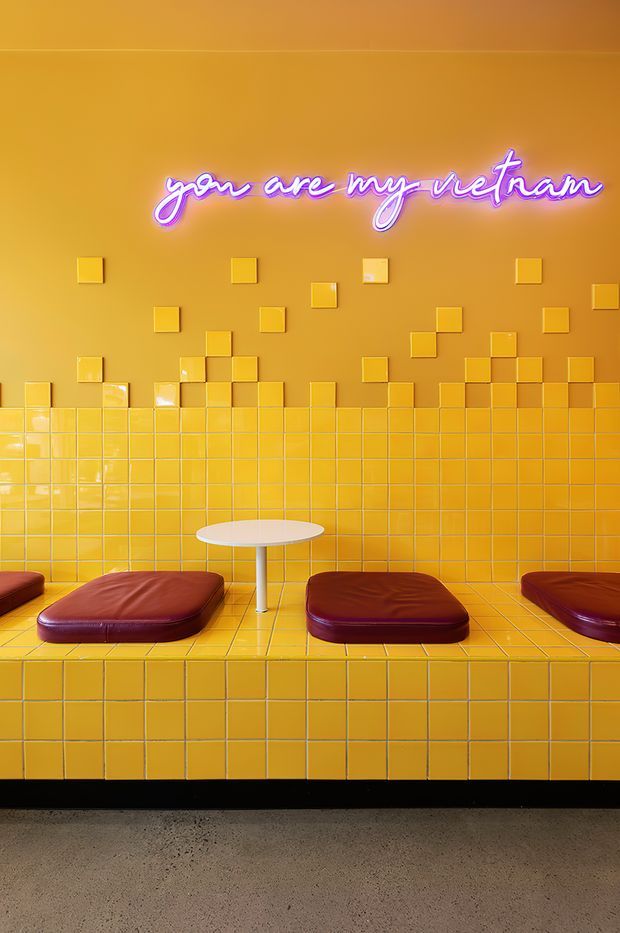A vivid, highly stylised interior seating area dominated by glossy mustard-yellow ceramic tiles covering the wall, bench platform, and front face. Three low burgundy leather cushions sit on the tiled bench, with a small white oval pedestal table between them. Scattered raised yellow square tiles create a geometric wall pattern, while a purple-pink neon script sign reads “you are my vietnam.” The space has a playful retro diner or hospitality atmosphere with strong colour contrast and polished surfaces.