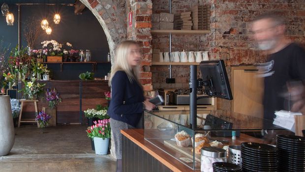 A rustic cafe or retail flower shop interior with an exposed red-brick arched opening, rough brick walls, dark painted surfaces, timber cabinetry, pendant lighting, and a glass-fronted service counter. Potted flowers and floral arrangements occupy the rear area, while a point-of-sale station and stacked tableware define the foreground. The image has an industrial, adaptive-reuse character with warm, atmospheric lighting.