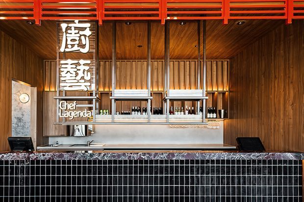 A dramatic commercial bar or restaurant counter with dark timber wall and ceiling lining, a black grid-tiled counter front, polished stone bar top, stainless-steel service shelving, and vivid red horizontal louvres or grilles at the ceiling. Vertical illuminated signage and bar equipment reinforce the hospitality setting.