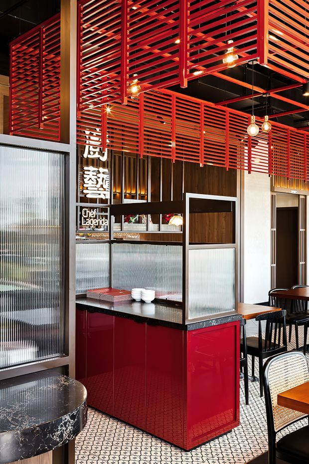Interior view of a stylised Asian-inspired restaurant dining space, featuring a glossy red service counter, patterned ceramic tile flooring, ribbed translucent glass partitions, dark timber wall panelling, dramatic red horizontal timber screens and a black exposed ceiling. Pendant globe lights, dining tables and black-framed cane chairs complete the contemporary hospitality fit-out. Chinese characters and restaurant signage are visible in the background.
