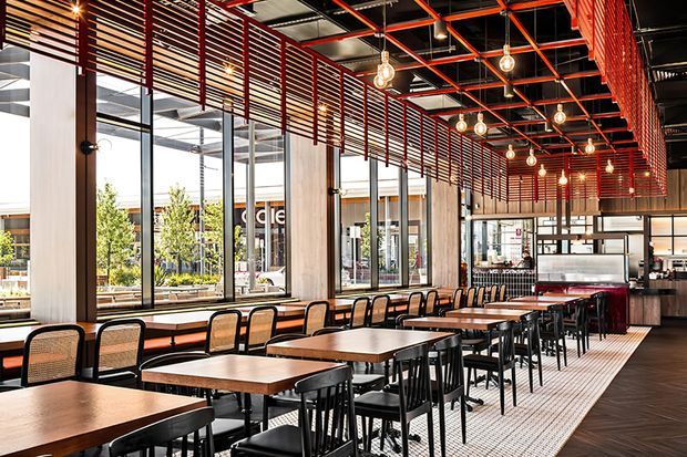 A spacious contemporary commercial dining area, likely a restaurant or food hall, with rows of timber tables and mixed chairs arranged beside floor-to-ceiling glazing. The interior features exposed concrete columns, a dramatic red exposed ceiling grid, horizontal red timber screening, black-framed windows, pendant globe lighting, and a service counter at the rear. The space is bright, open, and industrial-inspired.