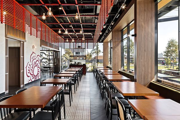A bright, modern commercial dining space with rows of wooden tables and black chairs, large windows along one side, a patterned tile walkway, timber wall panelling, and prominent red slatted ceiling and screening elements. Exposed black ceiling services, pendant lighting, and a graphic wall mural contribute an industrial yet warm restaurant interior.