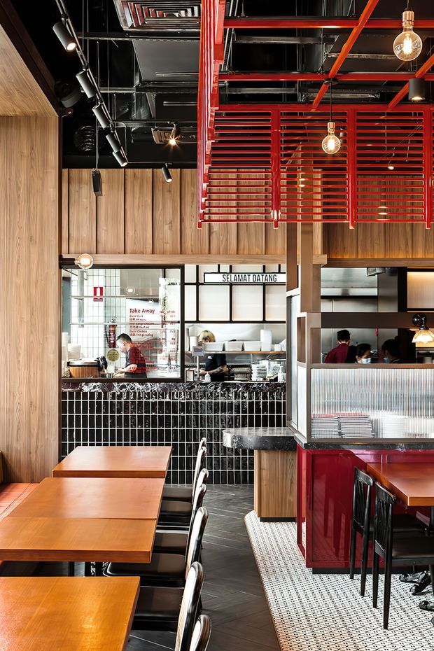 Interior view of a restaurant or casual dining venue with a strong industrial-inspired fit-out. Warm timber wall panelling and dining tables contrast with black tiled walls, black exposed services and ceilings, vivid red steel screens and joinery, pendant globe lights, and black metal-framed furniture. The image looks toward a service counter and open kitchen, with dining tables and booths in the foreground. Visible signage includes “SELAMAT DATANG” and takeaway information, reinforcing the hospitality setting.