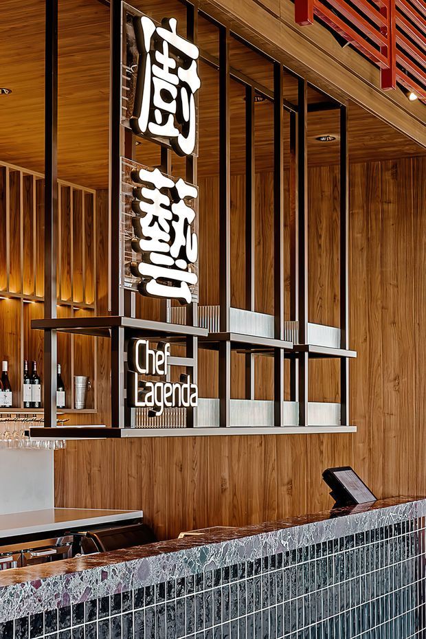 Interior view of a refined restaurant or bar reception counter, featuring extensive warm timber wall and ceiling panelling, black metal-framed open shelving, illuminated sculptural signage, wine and glass storage, and a tiled front counter with a distinctive mottled stone benchtop. The space has a polished hospitality fit-out with strong vertical detailing and integrated warm lighting.