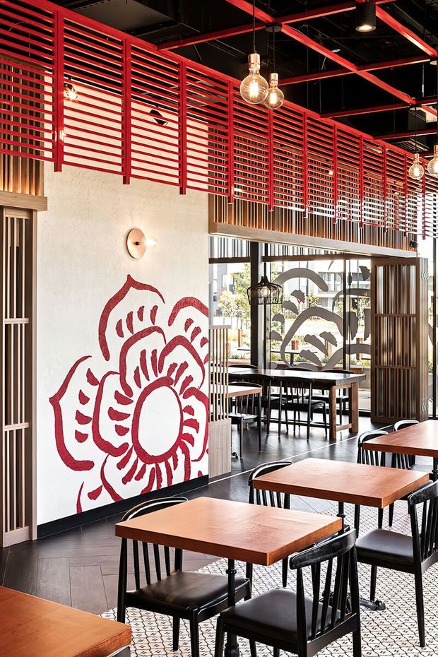 A stylish commercial dining interior, likely a restaurant or café, with timber tables, black chairs, patterned tiled flooring, and a large hand-painted floral mural. Strong red horizontal slatted screens and exposed ceiling beams create a bold graphic feature, complemented by warm pendant and wall lighting, dark ceiling finishes, and extensive glazing with decorative graphics.