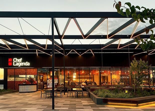 A contemporary outdoor dining plaza in front of a restaurant branded “Chef Lagenda.” The commercial facade features dark cladding, extensive floor-to-ceiling glazing, warm interior lighting, and a dramatic exposed steel canopy structure with triangular bracing. Outdoor tables and chairs sit on patterned paving beside raised landscaped garden beds and integrated bench seating.