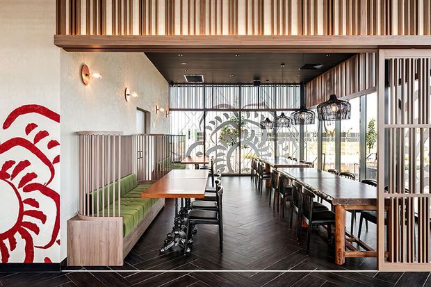A contemporary restaurant or café dining interior with long communal tables, upholstered green banquette seating, black dining chairs, pendant lanterns, and large glazed openings. Warm timber slat screens and ceiling detailing create a strong architectural rhythm, while a bold red-and-white mural occupies the left wall. The dark herringbone floor and black ceiling contrast with the warm timber and abundant daylight.