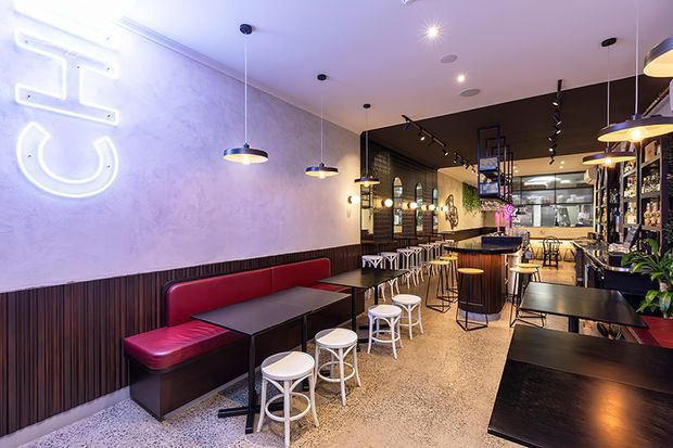 A narrow contemporary hospitality interior, likely a bar or casual restaurant, viewed along a long seating area toward the service counter. The space features red upholstered banquette seating, black tables, numerous white and yellow stools, dark timber wall panelling, a textured light wall, terrazzo-style flooring, pendant lights, track lighting, and a back bar with shelving and bottles. A neon wall sign and warm accent lighting add visual energy.