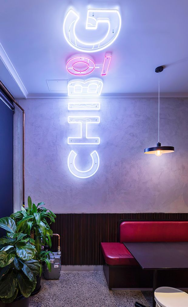 A moody commercial-style dining or lounge interior featuring a textured plaster wall with vertically arranged white and pink neon signage, a black pendant light, dark fluted wall panelling, a red upholstered booth, a dark table, exposed copper pipework, terrazzo flooring, and a large leafy plant. The space has a bold retro-industrial atmosphere with strong coloured lighting.