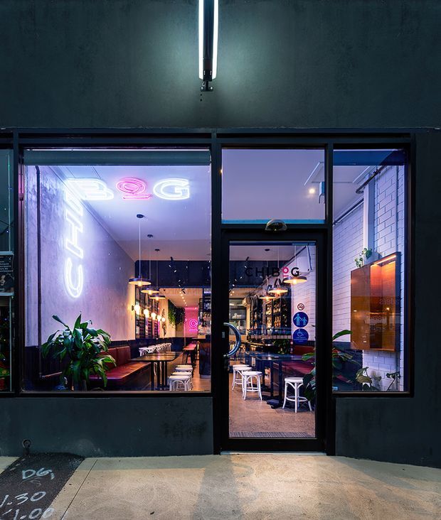 Night-time exterior view of a narrow contemporary commercial venue, likely a café or restaurant, seen through a dark-painted façade with large black-framed windows and a glazed entry door. The interior features neon signage, pendant lighting, bar or counter seating, stools, tables, plants, and a white brick wall, creating a vibrant urban hospitality atmosphere.