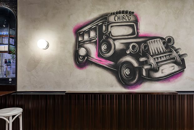 A stylised interior feature wall with a large black-and-white vintage bus mural accented in vivid pink. The wall has a textured grey plaster finish, a round wall light, dark vertically fluted timber panelling along the lower section, and a partial bar-height stool at the left. An arched glazed opening or mirror is visible at the far left.