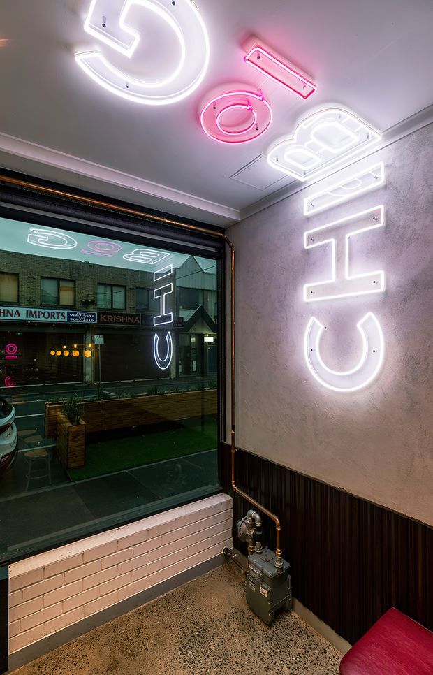 A compact commercial interior corner with a large street-facing window, exposed gas piping and meter, dark fluted lower wall panelling, textured plaster walls, terrazzo-style flooring, and prominent white and pink neon lettering and symbols mounted on the walls and ceiling. Reflections in the glazing show the neon signage and the street-facing storefront opposite. A red upholstered seat is partially visible in the foreground.