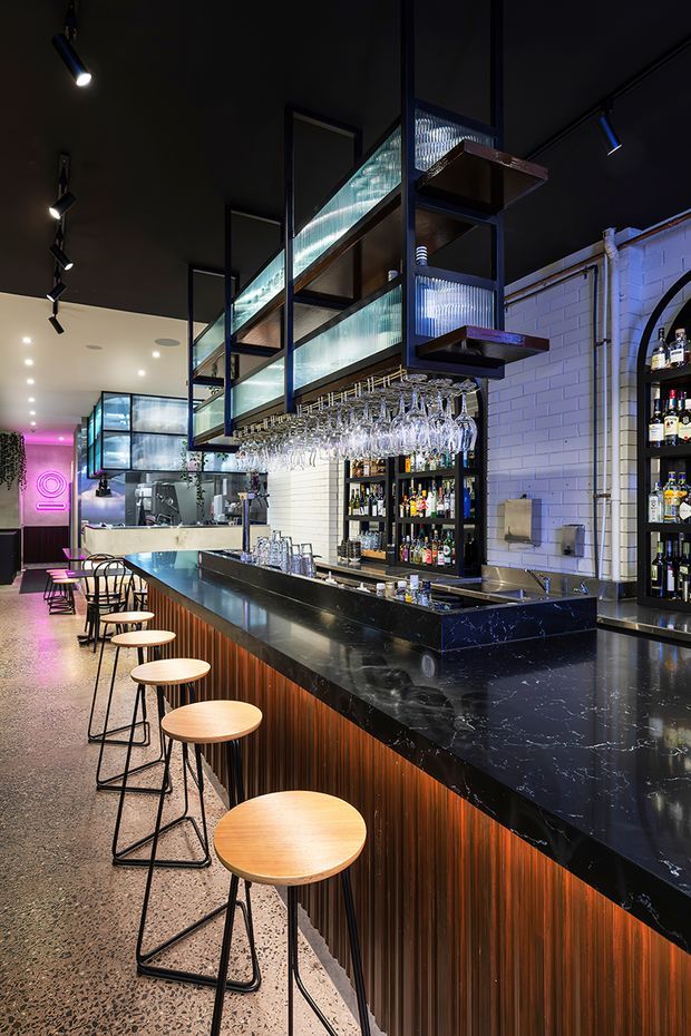A moody contemporary commercial bar interior with a long black stone bar counter, warm vertically ribbed timber front, and a row of wooden stools with black metal frames. Suspended open shelving with translucent ribbed glass panels displays glassware above the bar, while back-bar shelving holds bottles and equipment. White glazed brick walls, black ceilings, track lighting, polished concrete or terrazzo flooring, and pink accent lighting create a refined hospitality atmosphere.