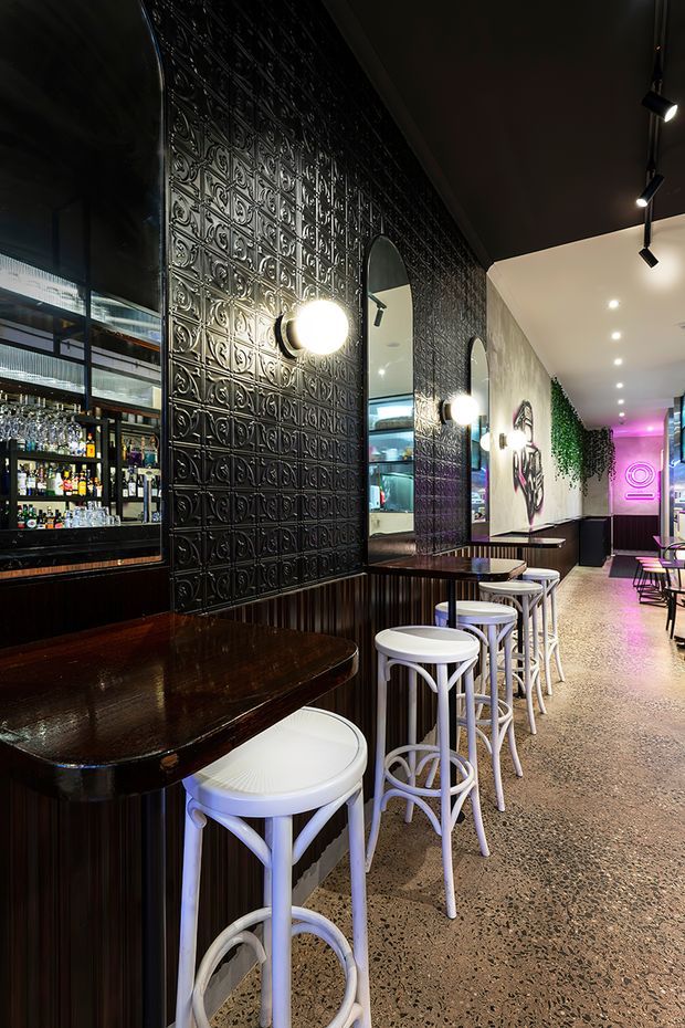 A narrow, stylish bar or hospitality venue with a long dark timber counter and matching built-in seating, white bentwood bar stools, mirrored panels, and a black embossed metal feature wall. The space includes a backlit bottle display, track lighting, arched mirrors, terrazzo flooring, wall artwork, greenery, and vivid pink neon lighting toward the rear. The overall atmosphere is moody, urban, and eclectic.