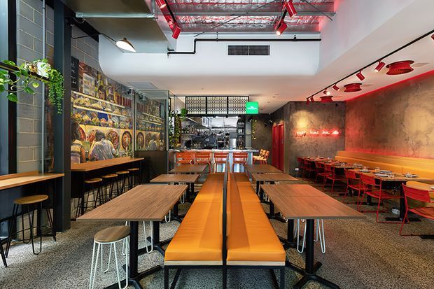 A modern industrial-style restaurant or café interior with long communal tables, orange upholstered banquette seating, red chairs, bar seating, and exposed services. The space features a terrazzo-like speckled floor, dark wall finishes, large illustrated wall graphics, concrete-look surfaces, red-painted ceiling pipes and ductwork, track lighting, and a partially visible service counter at the rear.