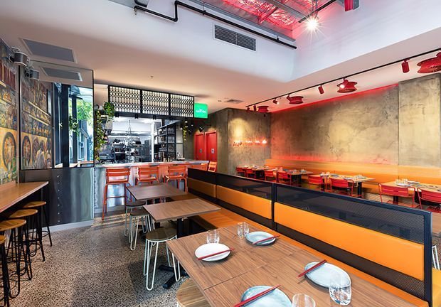 A vibrant industrial-style restaurant interior with rows of timber tables, orange upholstered bench seating, metal-framed chairs, and a bar or service counter at the rear. The space features polished concrete flooring, dark metal partitions, exposed ceiling services, track lighting, textured concrete walls, red accent lighting, and a partially visible commercial kitchen. A large colourful wall mural and street-facing glazing add visual interest.
