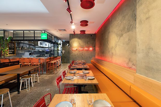 A contemporary restaurant interior with a long banquette dining area, timber tables, red metal chairs, and a visible open bar and kitchen counter. The space features a textured concrete-look wall, dramatic red linear lighting, exposed ceiling services, patterned screening, and terrazzo-style flooring.