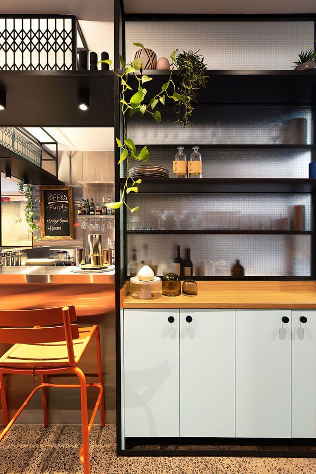 A compact, stylish hospitality bar or café interior with a black-framed glazed shelving partition, warm timber countertop, white lower cabinetry, terrazzo flooring, and vivid orange seating. The open bar area includes drinks shelving, pendant and recessed lighting, decorative plants, and a visible chalkboard menu. The design combines strong black metal detailing with warm timber and colourful furniture in a lively, refined setting.