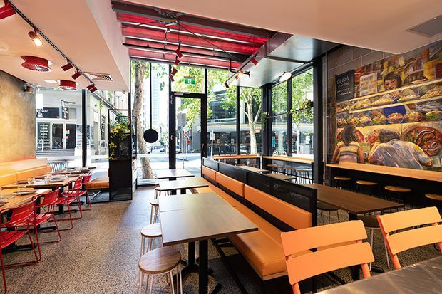 A contemporary commercial dining space, likely a café or casual restaurant, with extensive street-facing glazing, black-framed doors and windows, long upholstered banquette seating, timber tables, stools, and red and orange chairs. The interior features polished terrazzo-style flooring, exposed dark ceiling services, strong red ceiling accents, warm pendant lighting, and a large colourful mural along the right wall.