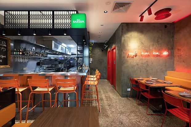 A stylish Asian-inspired restaurant interior with a bar and open kitchen on the left, counter seating in the foreground, and dining tables with orange banquette seating on the right. The space features a dark textured concrete wall, red doors and accents, patterned metal screens, terrazzo-style flooring, exposed services, and a red neon sign, creating a lively industrial atmosphere.