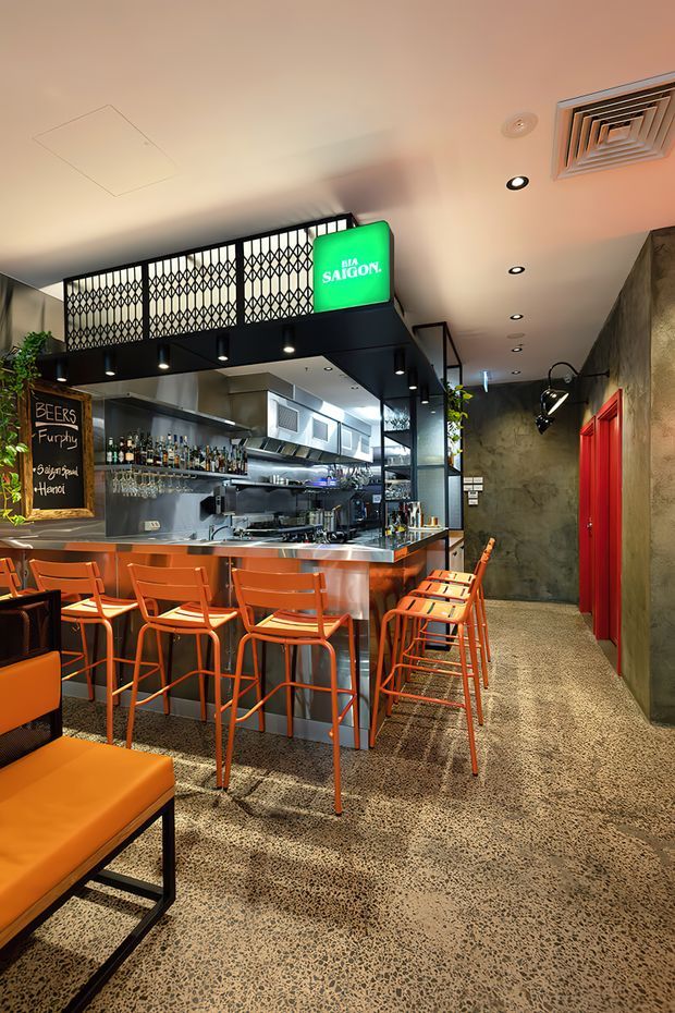 A compact contemporary bar and restaurant interior with a long polished metal service counter, open commercial kitchen, liquor display, orange bar stools and dining chairs, and a black overhead partition with decorative metal grilles. The space has a dark textured concrete wall, bright red doors, polished terrazzo flooring, exposed-style track and recessed lighting, and a prominent green Saigon beer sign. The atmosphere is urban, energetic, and industrial-inspired.