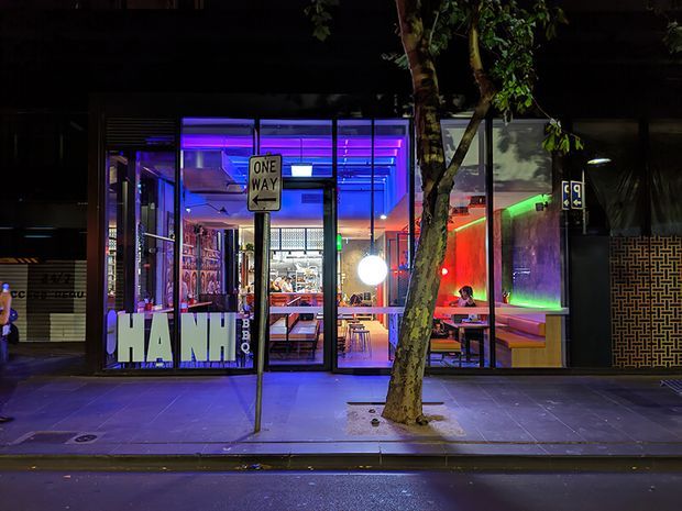 Night-time street view of a brightly illuminated urban hospitality or commercial interior behind full-height glass storefront glazing. Strong blue, purple, red and green feature lighting illuminates visible tables, seating and wall surfaces inside. A tree, sidewalk, road, one-way sign and brick-patterned exterior wall frame the facade.