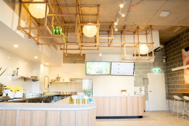 Bright contemporary café interior with a curved light-wood service counter, white tiled food-preparation area, pale timber cabinetry, exposed ceiling structure, spherical pendant lights, wall-mounted screens, and bar seating. A dark masonry feature wall and graphic artwork add contrast.