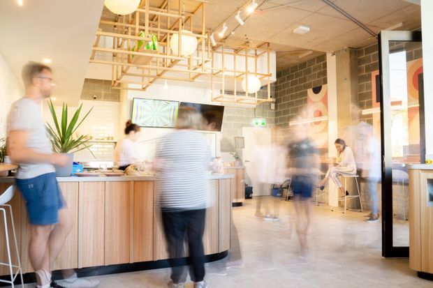 Bright contemporary commercial interior resembling a café or casual dining venue, with a central timber-clad service counter, exposed timber ceiling framework, pendant globe lights, concrete block walls, tiled flooring, and people moving through the space with motion blur. Large glazing and graphic wall artwork add visual interest.