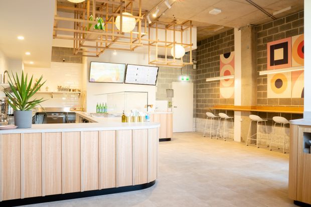 Bright contemporary commercial café or hospitality interior with a curved light timber service counter, pale benchtop, open kitchen and display area, exposed dark masonry block wall, timber-framed ceiling light installation, and a long high bar with white stools. Colourful geometric artwork adds a bold accent to the otherwise light, warm space.