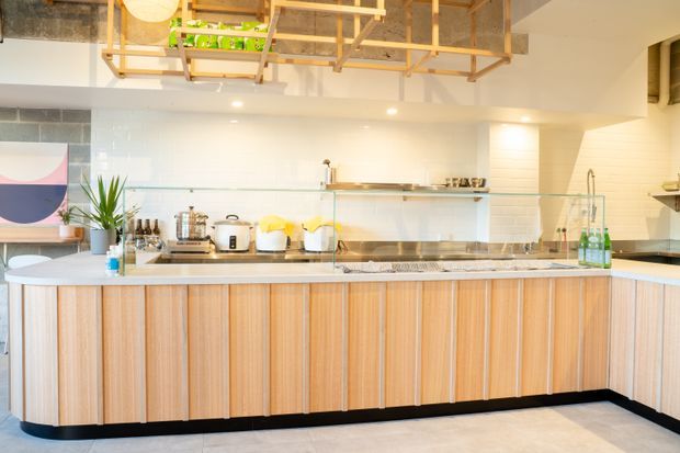Bright contemporary kitchen with a large island featuring vertically fluted light-wood cabinetry, a pale stone benchtop, white tiled walls, open shelving, stainless-steel appliances, and a distinctive timber ceiling rack with integrated lighting and storage.