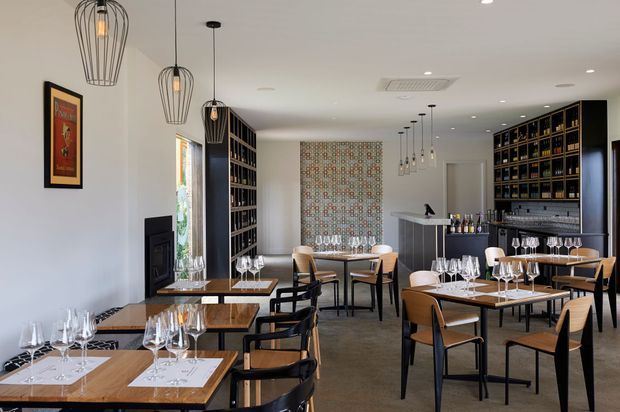 A refined contemporary restaurant or wine bar interior with multiple set dining tables, a central bar counter, extensive dark built-in wine shelving, a tiled backsplash, and a decorative patterned feature wall. Black wire pendant lights, timber tables, mixed timber-and-black chairs, and white walls create a polished hospitality setting.