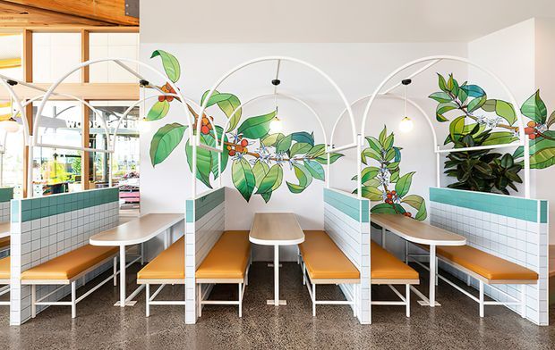 Bright contemporary café or commercial dining interior with repeated booth seating divided by white metal arched screens. The booth bases and walls are clad in small white square tiles with teal-green tile bands, paired with warm honey-toned timber tabletops and upholstered bench seating. A large botanical mural of leafy branches and berries spans the white wall, while pendant lights and extensive glazing brighten the space. Polished speckled concrete flooring and exposed timber ceiling elements are visible.