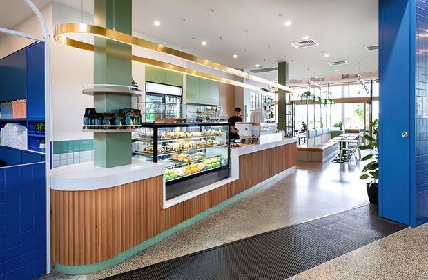 Bright, contemporary commercial café or food-service interior with a long timber-faced service counter, glass refrigerated display cases filled with prepared food, coffee equipment, shelving, and counter seating. The space features a polished terrazzo-style floor, teal-green tiled and painted surfaces, blue tiled columns and walls, extensive glazing, and distinctive linear brass pendant lighting. Large windows at the rear create an airy, welcoming atmosphere.