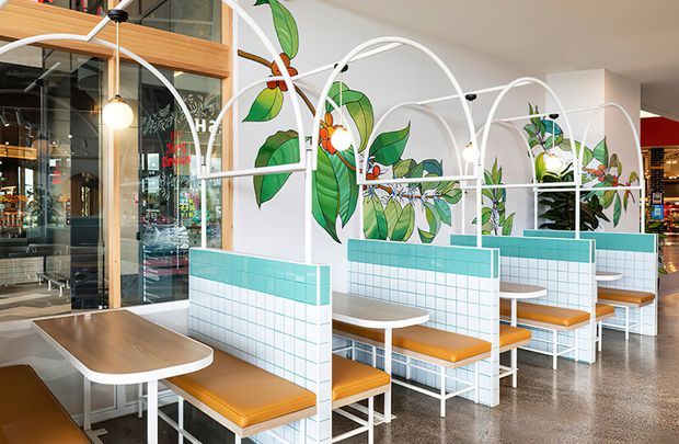 Bright contemporary commercial dining interior with repeated booth seating arranged behind low tiled partitions. White arched metal frames create a rhythmic canopy structure over turquoise-and-white grid-tiled dividers, with tan upholstered bench seats and light timber tables. Large tropical botanical murals add a vivid green accent, while polished concrete flooring, full-height glazing, and exposed timber framing contribute to a modern hospitality fit-out.