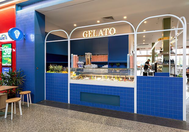 A brightly styled commercial gelato counter with deep blue tiled walls and cabinetry, white arched metal framing, a glass display case, illuminated GELATO signage, and colourful product shelving. A terrazzo-like floor, stools, plants, and adjacent retail elements contribute to a playful, contemporary food-service interior.