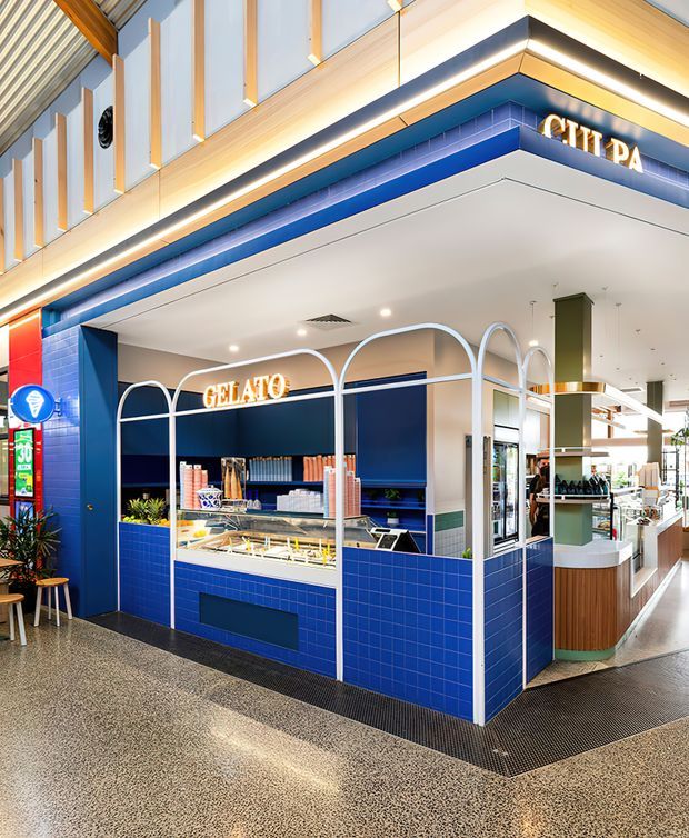 A bright contemporary gelato kiosk and café tenancy in a shopping-centre interior. The fit-out features a bold cobalt-blue tiled counter and walls, curved white metal-framed arches, illuminated signage, integrated gelato display cases, timber cabinetry, and a polished terrazzo-style floor. The wider retail environment and high-level architectural ceiling are visible around the kiosk.