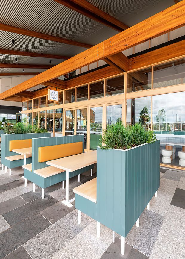 A bright contemporary commercial dining or hospitality space with outdoor-style booth seating, pale timber tables, teal tiled planter partitions, and mustard-yellow upholstered backrests. The space features extensive floor-to-ceiling glazing, exposed warm timber beams, a corrugated metal ceiling, and a large-format tiled floor. Planting-filled built-in planters separate the seating areas, while the glazing reveals a bright exterior setting and additional interior seating.